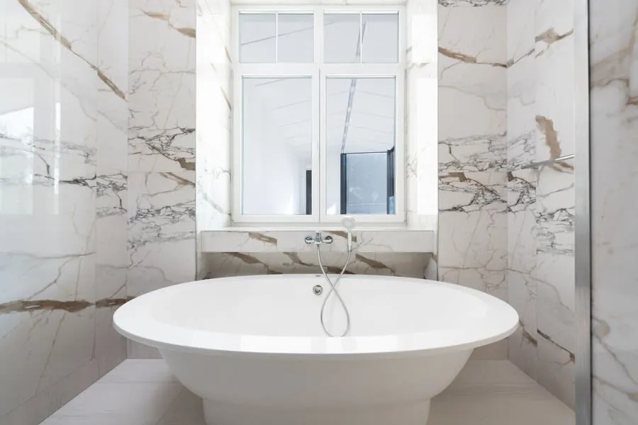Freestanding bath with clear space around it in a marble bathroom with a large window