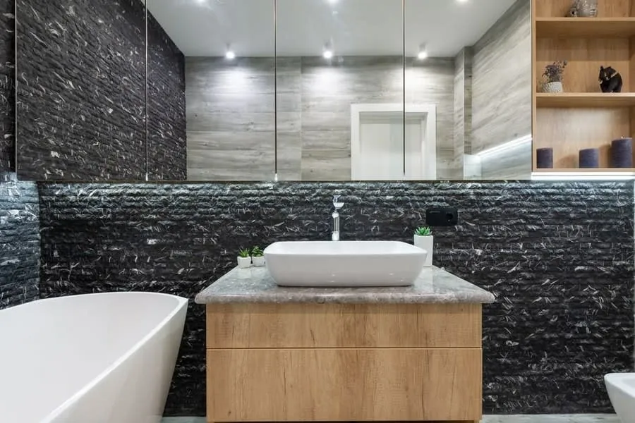 Freestanding bath beside an oak vanity and textured black stone wall